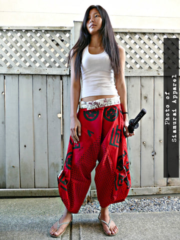 A person wearing red samurai pants with black details and a white top, standing against a wooden backdrop.